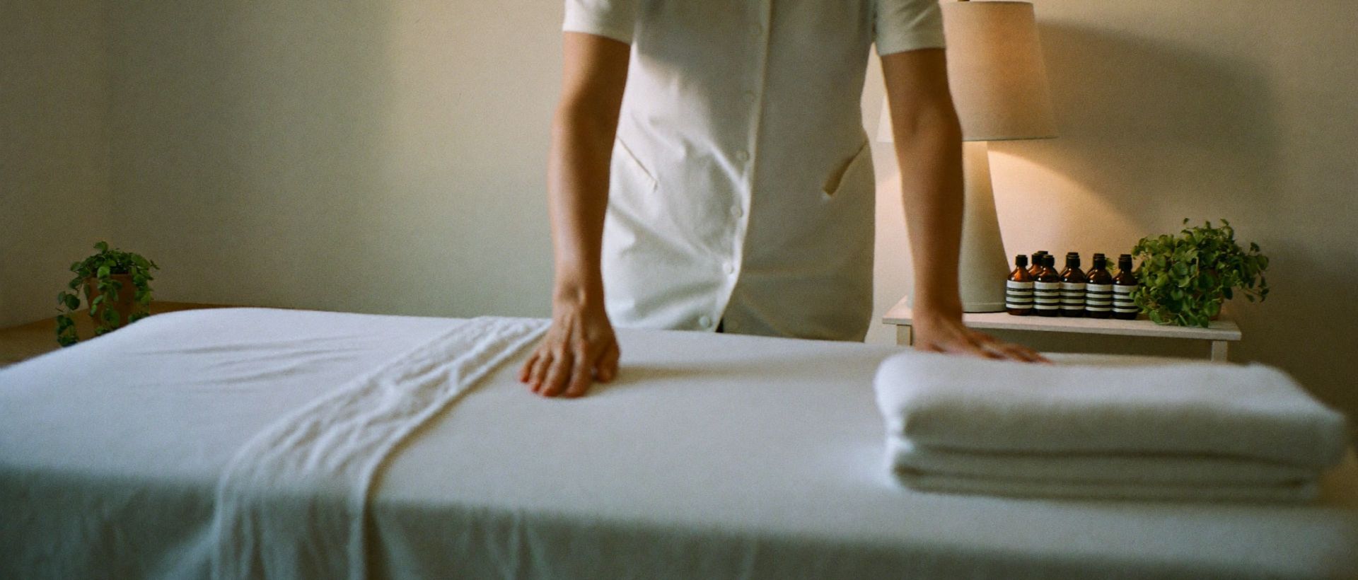 A beauty therapist preparing a treatment room with folded towels and warm lamp light