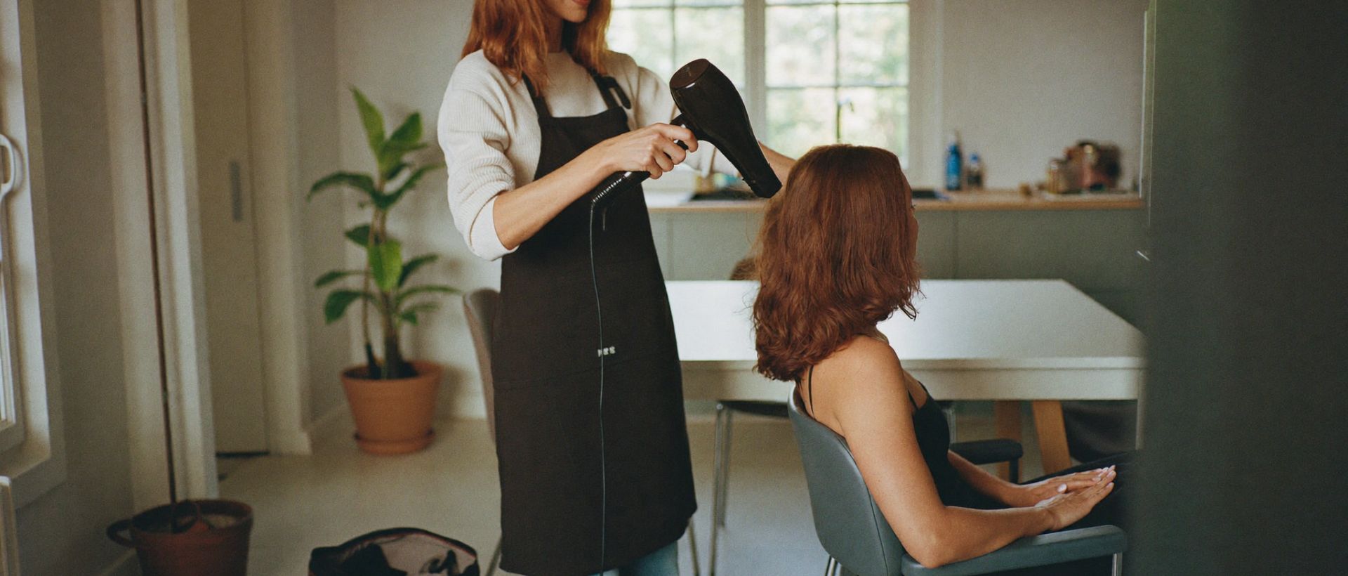 A mobile hairdresser styling a seated client's hair in a bright home kitchen